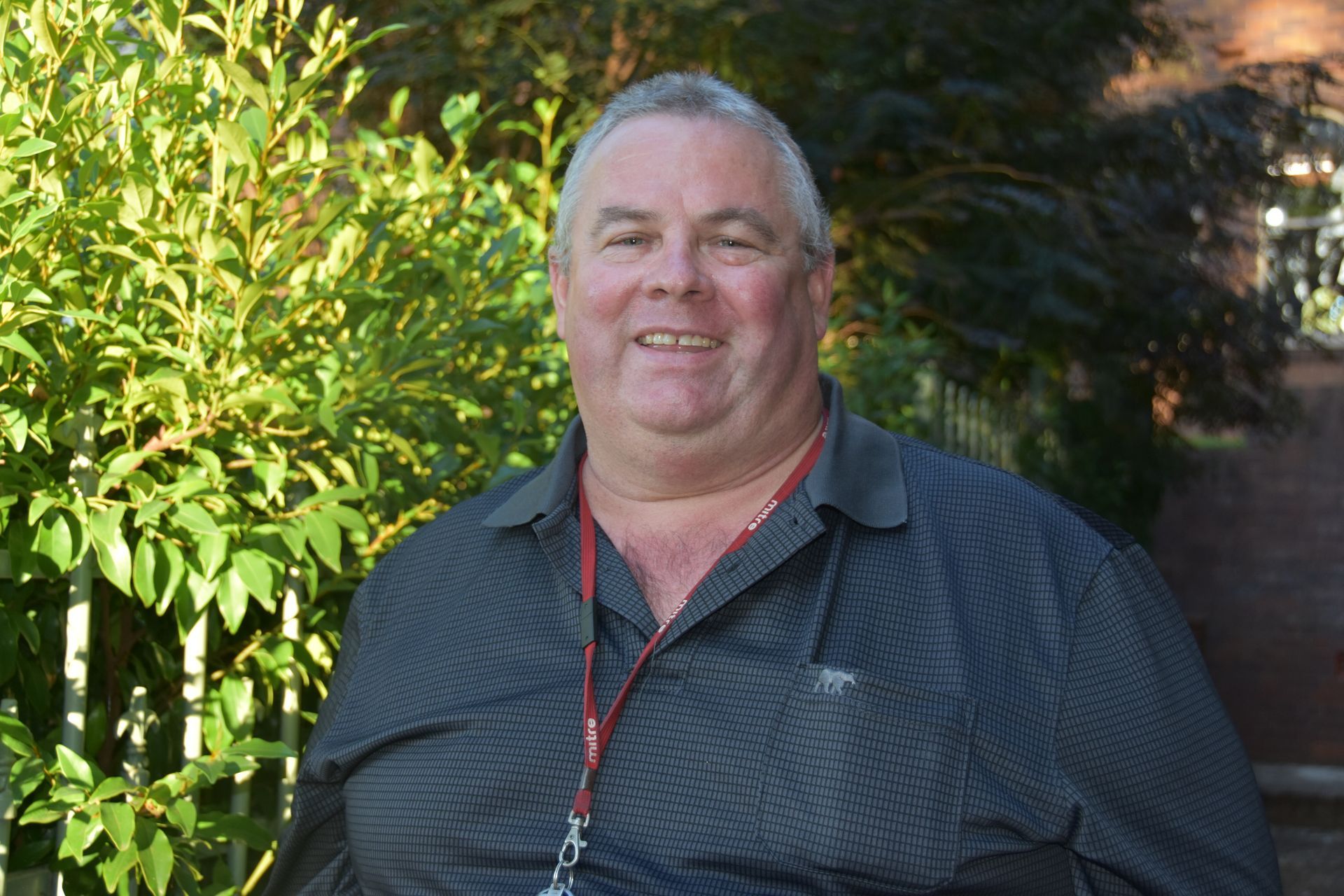 A man wearing a lanyard around his neck is standing in front of a bush.