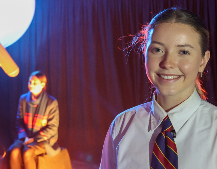 A student in a white shirt and tie is smiling in a dark room.