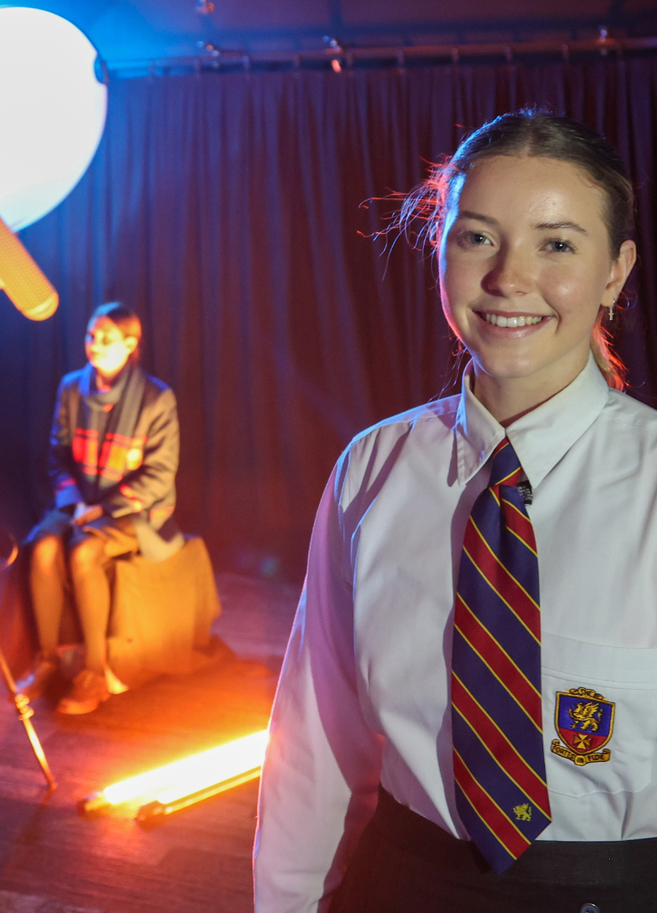 A student in a white shirt and tie is smiling in a dark room.