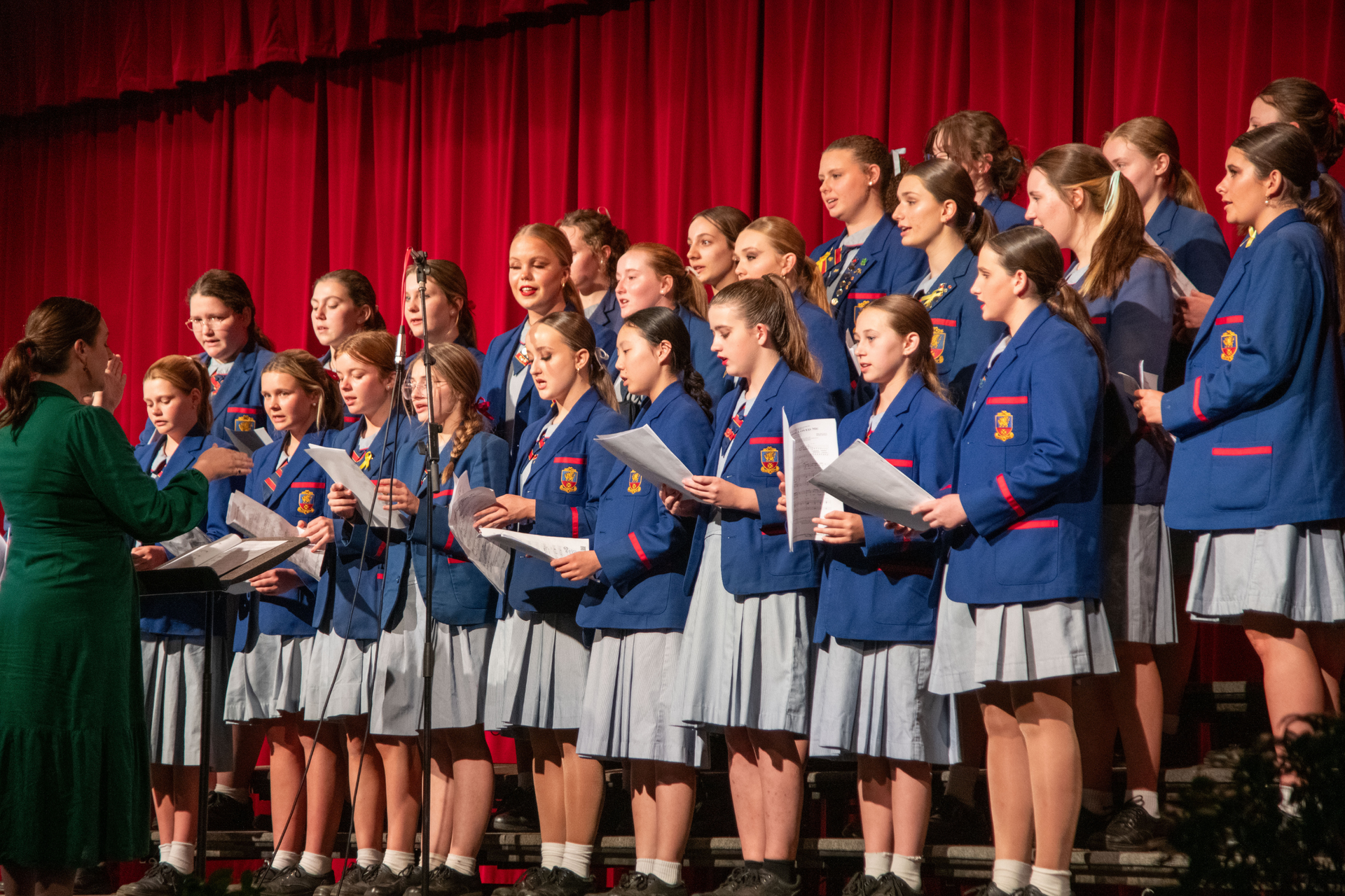 A choir of young students are singing on a stage in front of a red curtain.