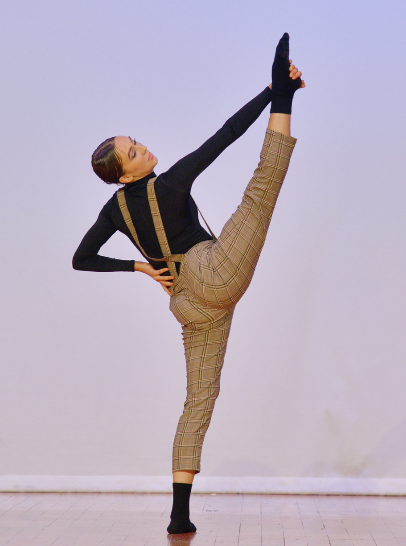 A student in suspenders is doing a split on a wooden floor.