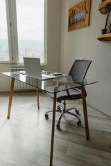 A glass-topped desk with a laptop and swivel chair in a sunlit room featuring a wall painting and floating wooden shelves.