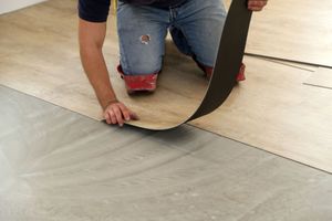 A person kneeling on a concrete floor installs light-colored wood-look vinyl plank flooring.