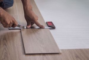 A person uses a utility knife and a metal square to score a wood-look vinyl plank during flooring installation.