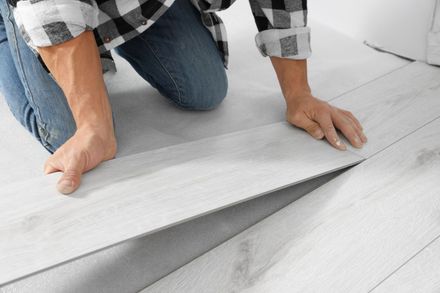 A person wearing a plaid shirt kneels on the floor, installing light-gray wood-look laminate flooring planks.