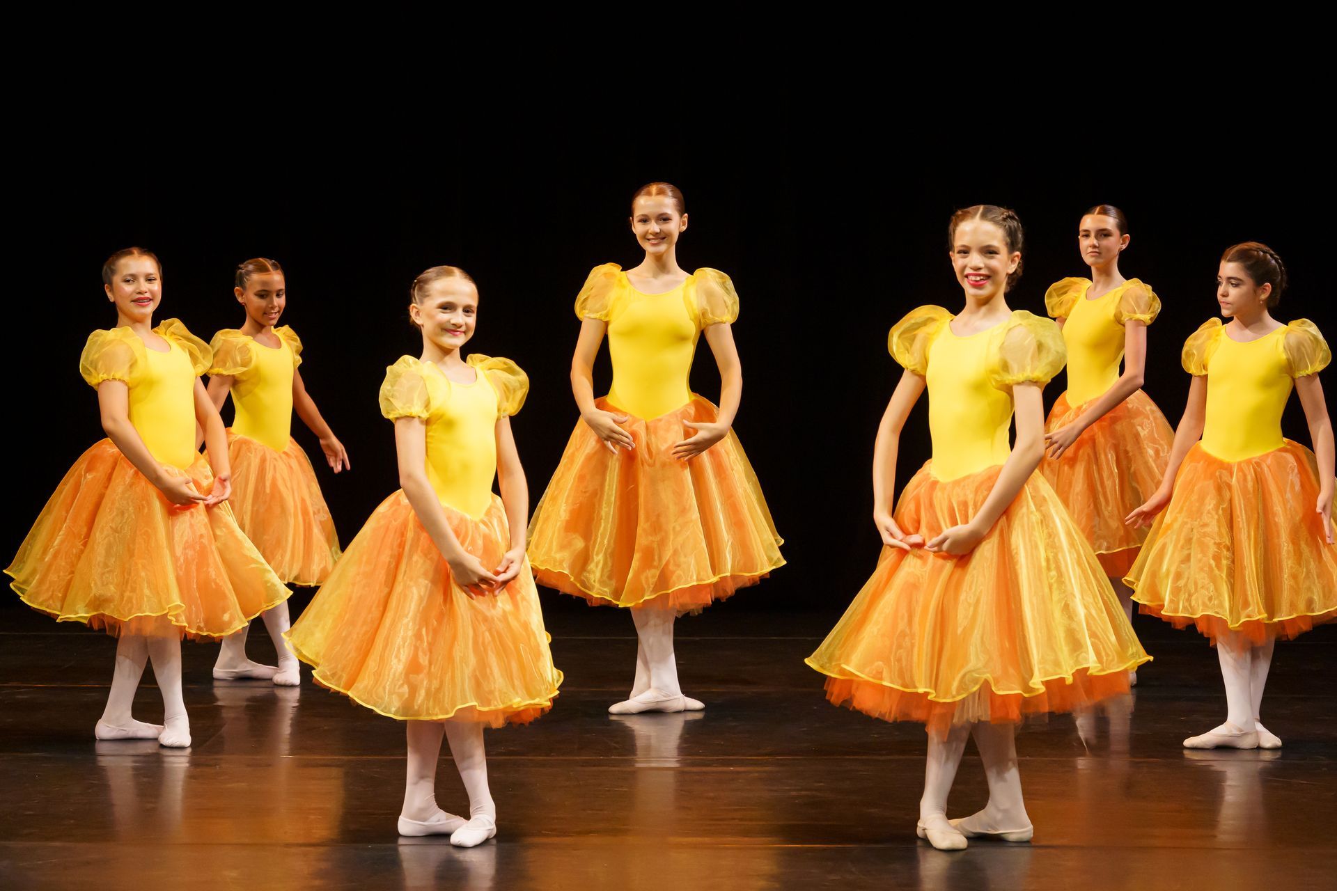 Bailarinas vestidas com tops amarelos e tutus laranja se apresentam em um palco.