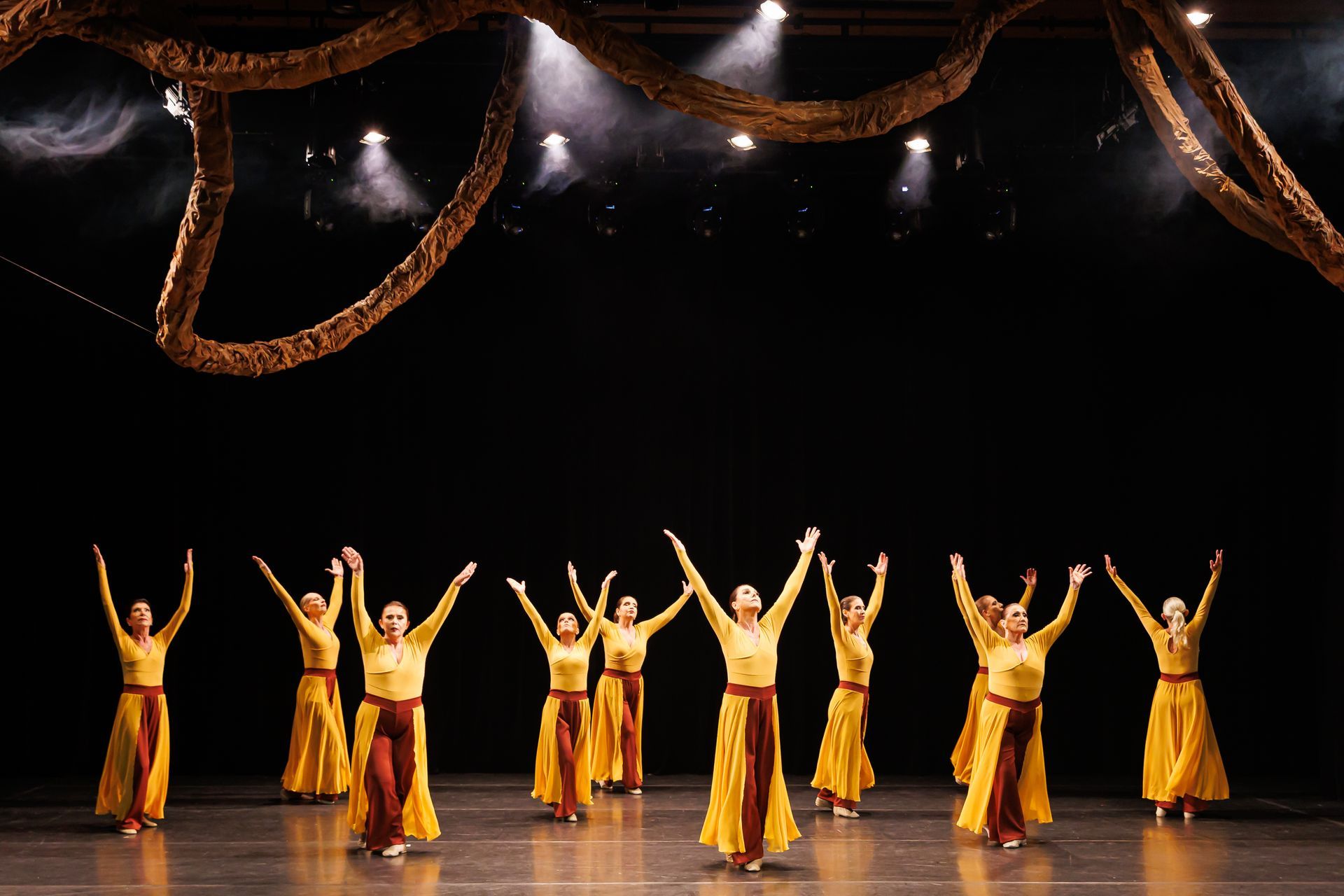 Um grupo de dançarinas com vestidos amarelos e marrons, com os braços erguidos, em um palco com um fundo escuro.