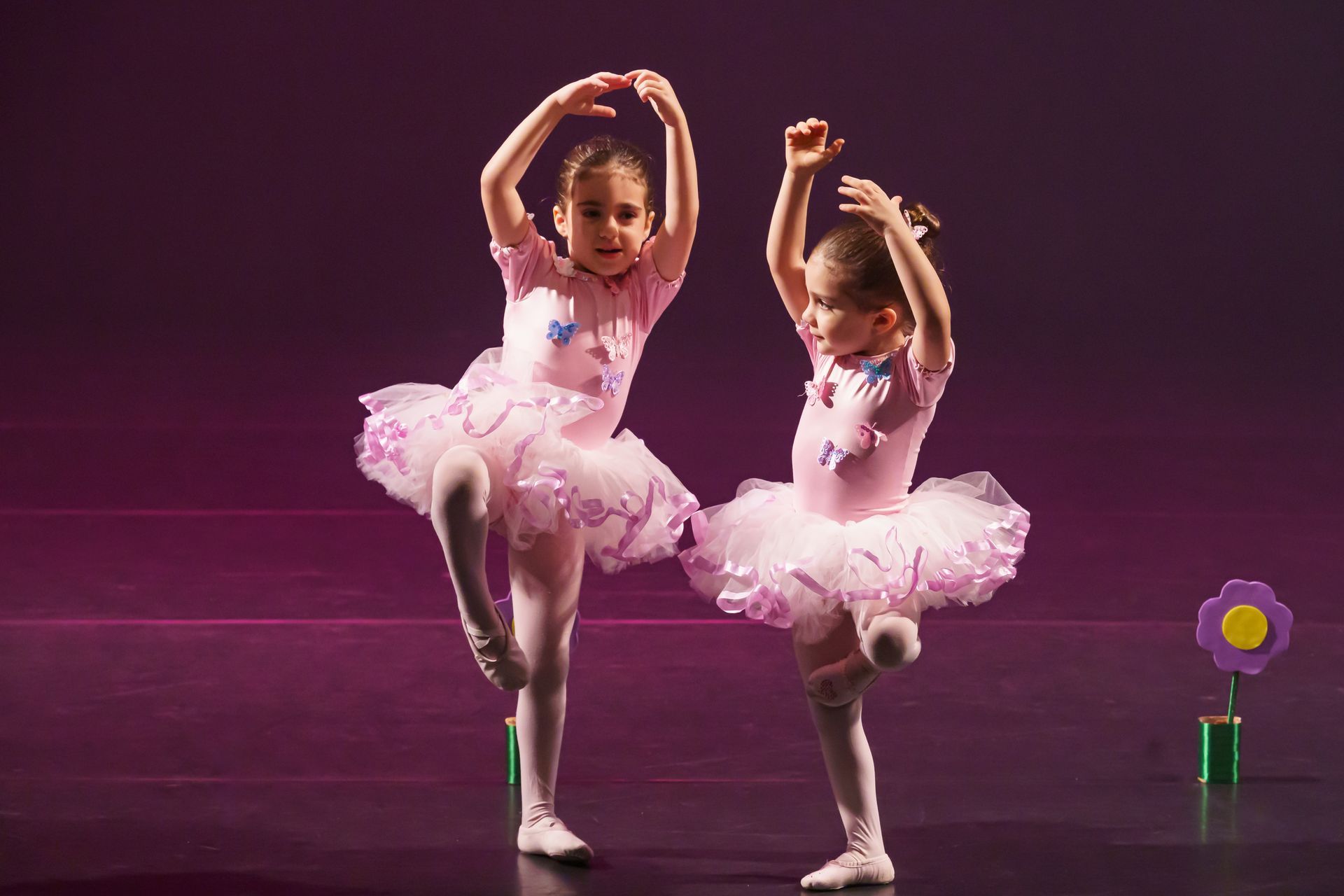 Duas jovens bailarinas de tutu rosa, equilibrando-se em uma perna só, com os braços erguidos, no palco.