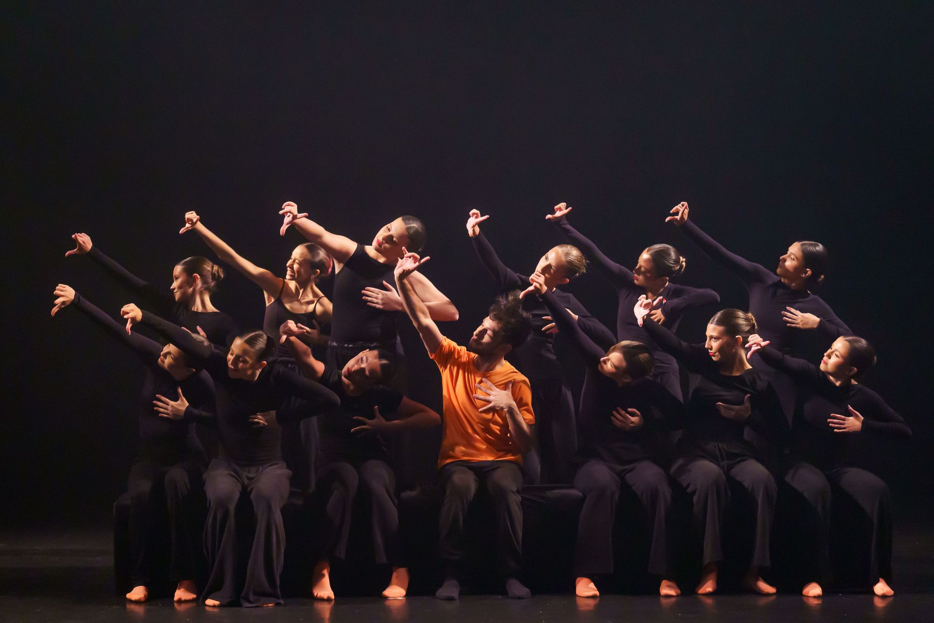 Dançarinas vestidas de preto, uma delas de laranja, posando com os braços erguidos em um palco escuro.