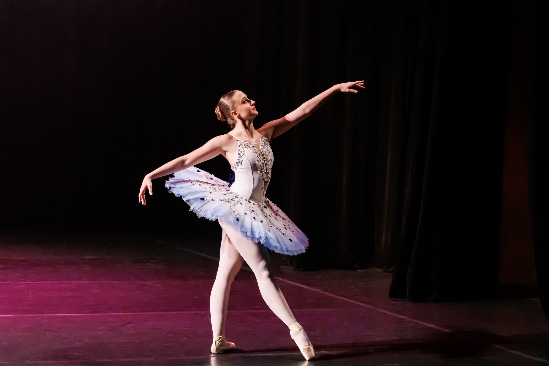 Bailarina de tutu branco e sapatilhas de ponta no palco, com os braços estendidos.