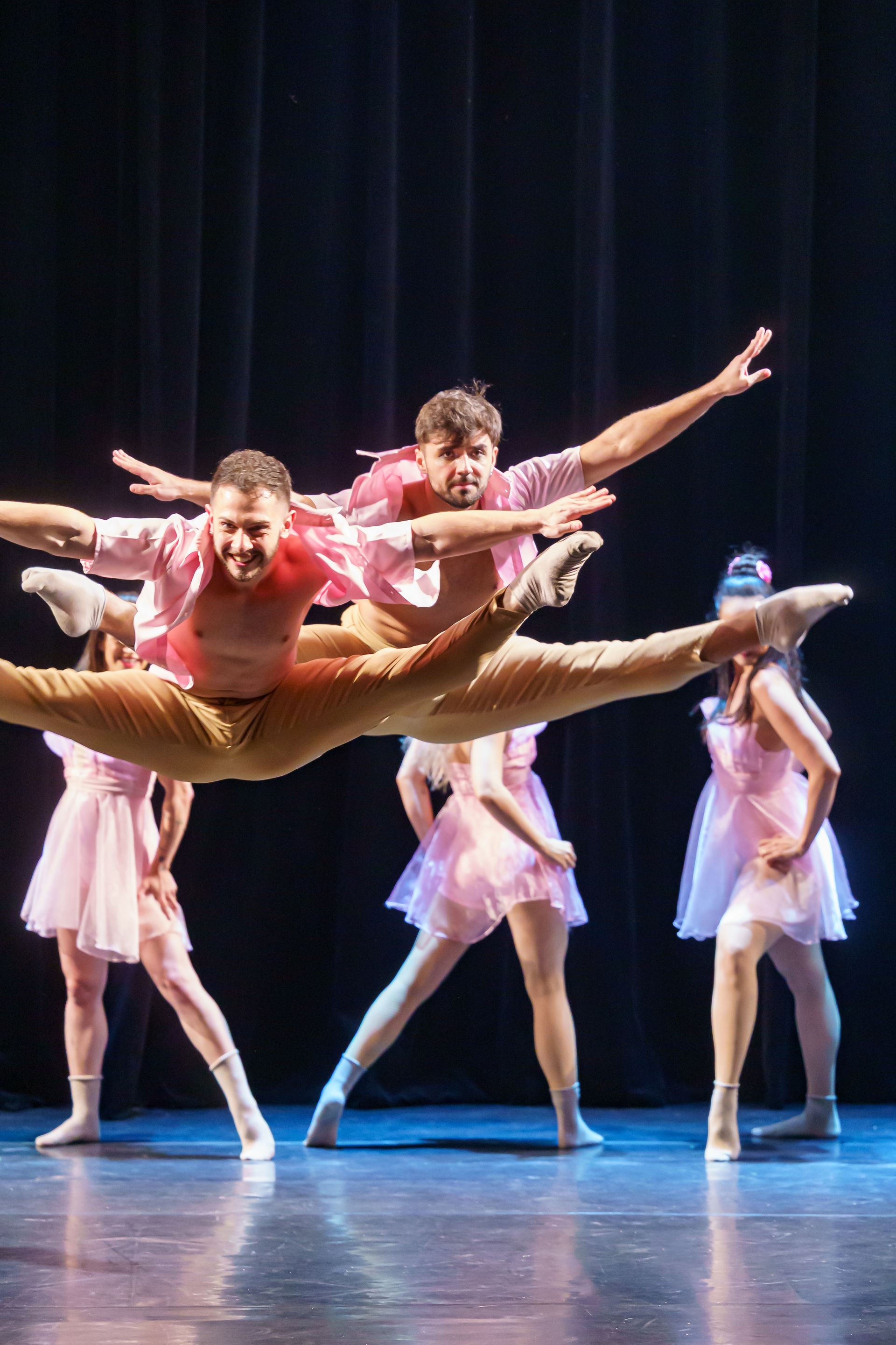 Dançarinos com trajes rosa e bege executam um salto com abertura de pernas no palco.