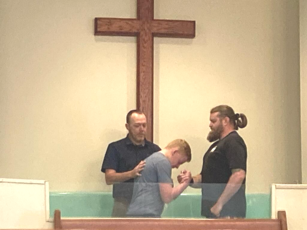Three men stand in a baptismal pool in front of a wooden cross; one man leans forward while the others support him.