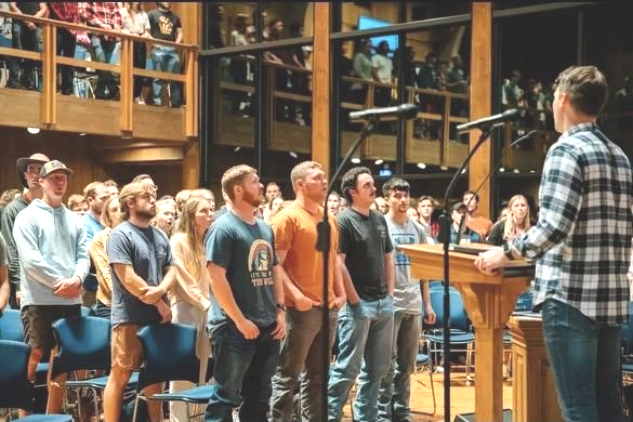 People stand in a wood-paneled hall, facing a person speaking at a podium with microphones, suggesting a church service.