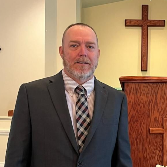 A person in a suit and plaid tie standing in front of a wooden pulpit with a cross on the wall behind them.