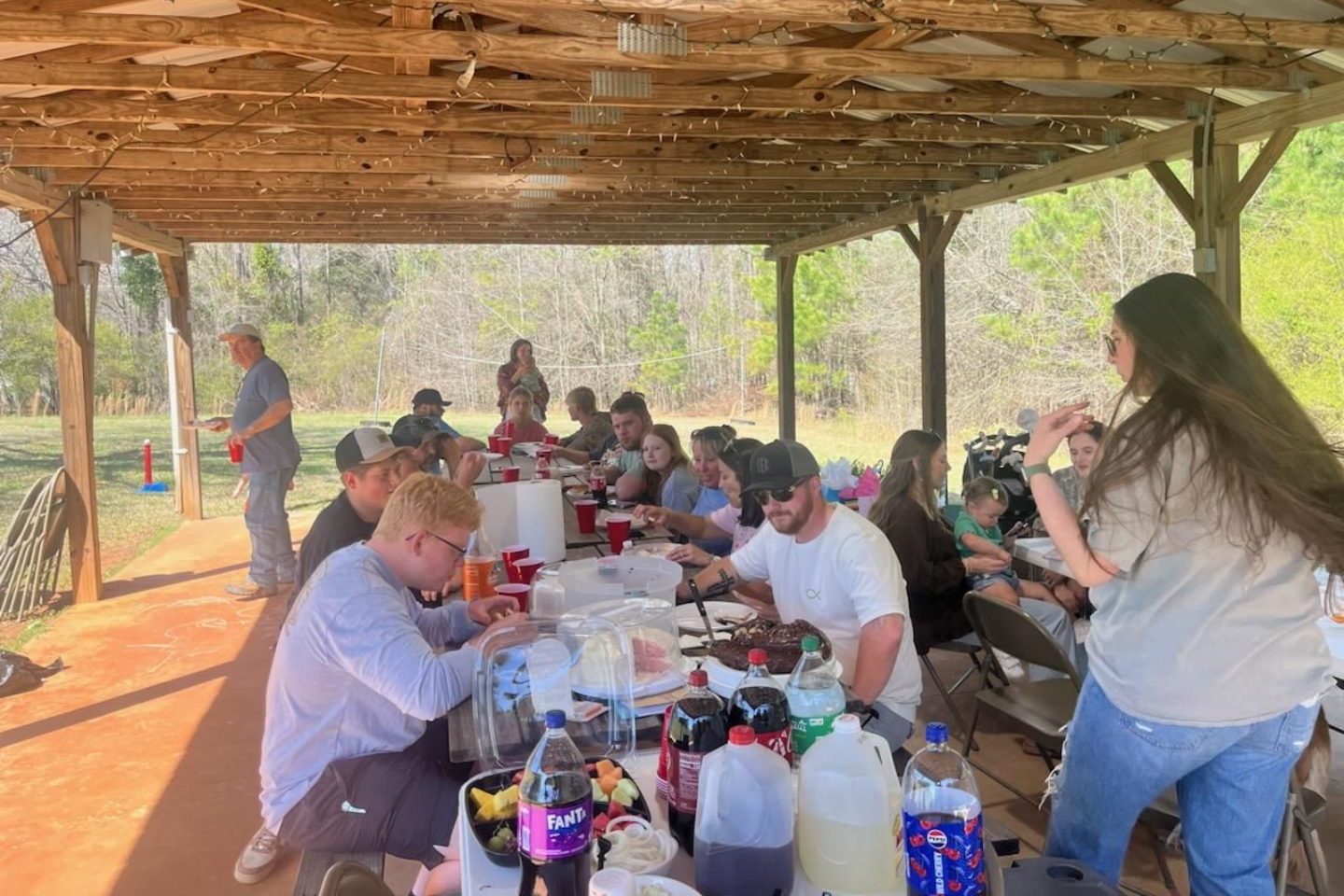 A group of people gathers under a rustic outdoor pavilion for a meal, seated at long tables with food and drinks.