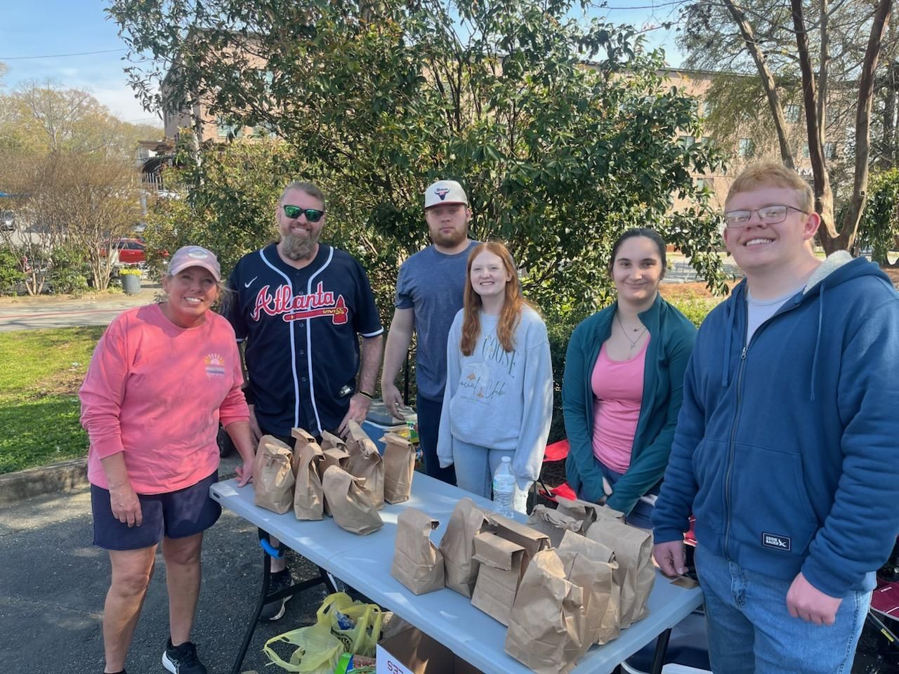 A group of six people stand behind a table filled with brown paper bags, outdoors in a park-like setting.