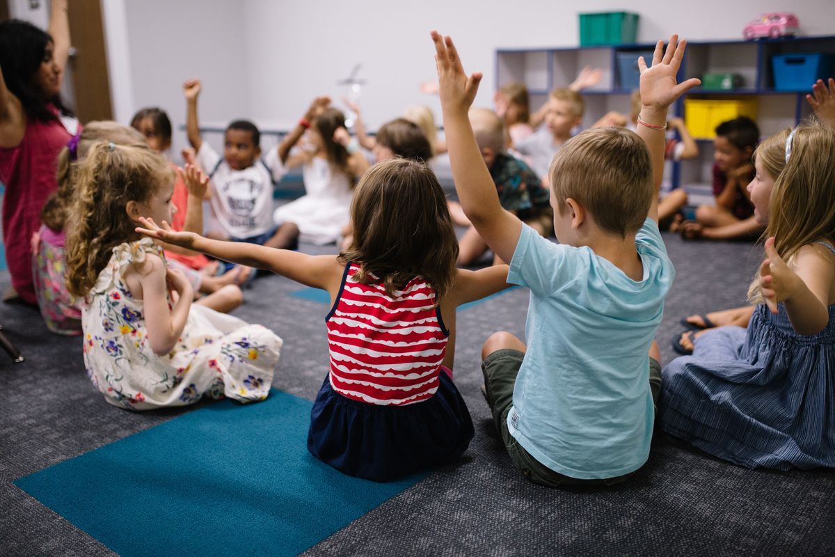 Children sitting in a circle on the floor with their hands raised in a classroom.