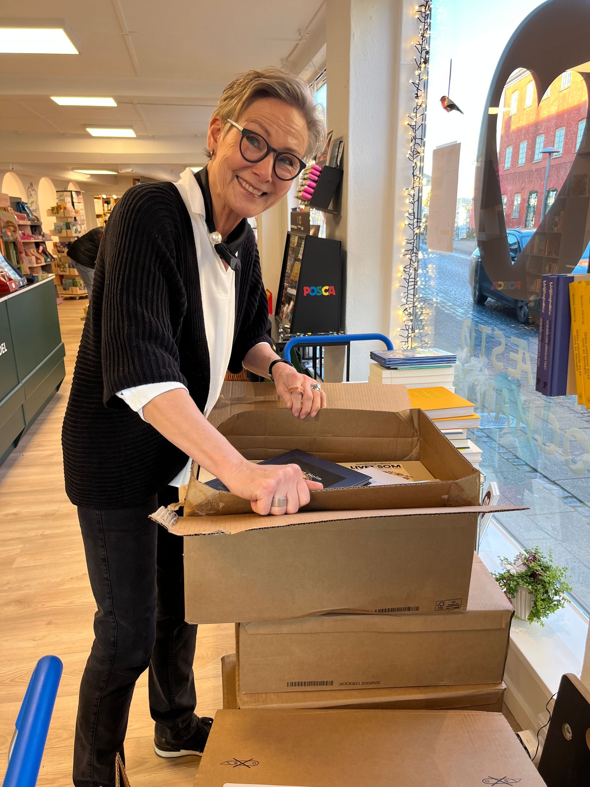 Kvinde smiler, mens hun pakker kasser ud i en butik. Hun har briller og en sort sweater på og står ved et vindue.