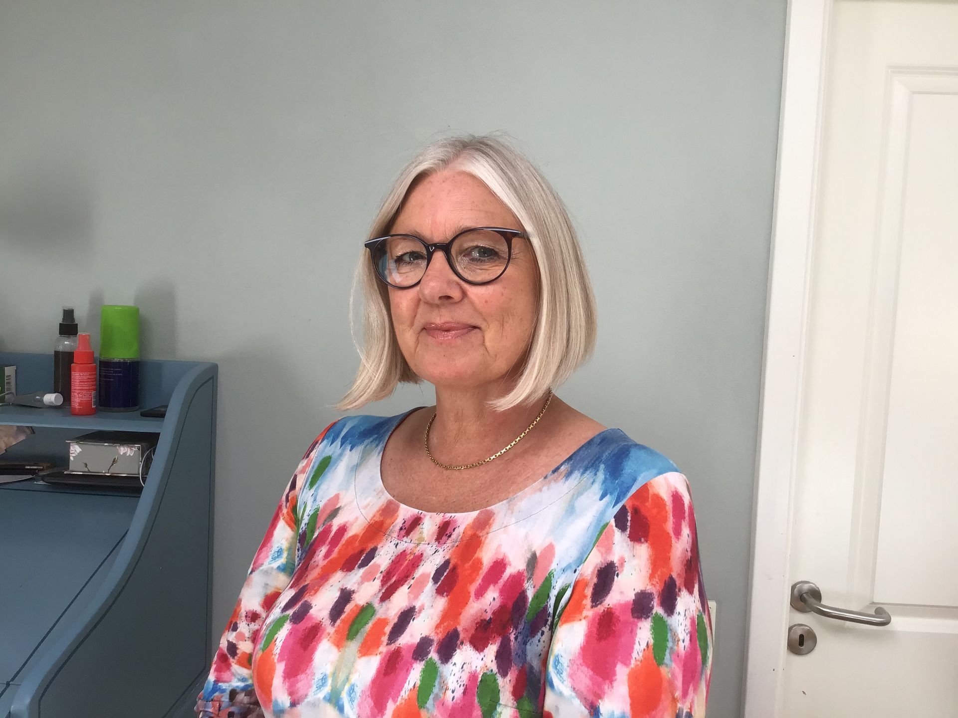 En kvinde med briller og en farverig skjorte står foran en dør.
