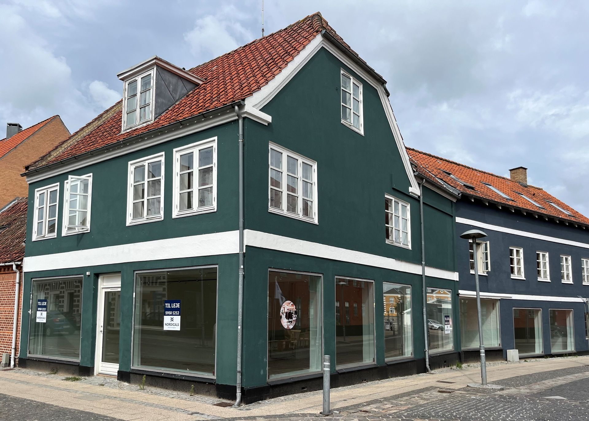 Grønne og blå bygninger med tomme butiksfacader og rødt tegltag. Bygningen ligger på en brostensbelagt gade.