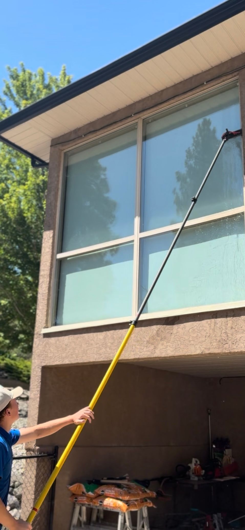 A person cleaning a window with a long yellow pole. The person is standing outside a brown building.