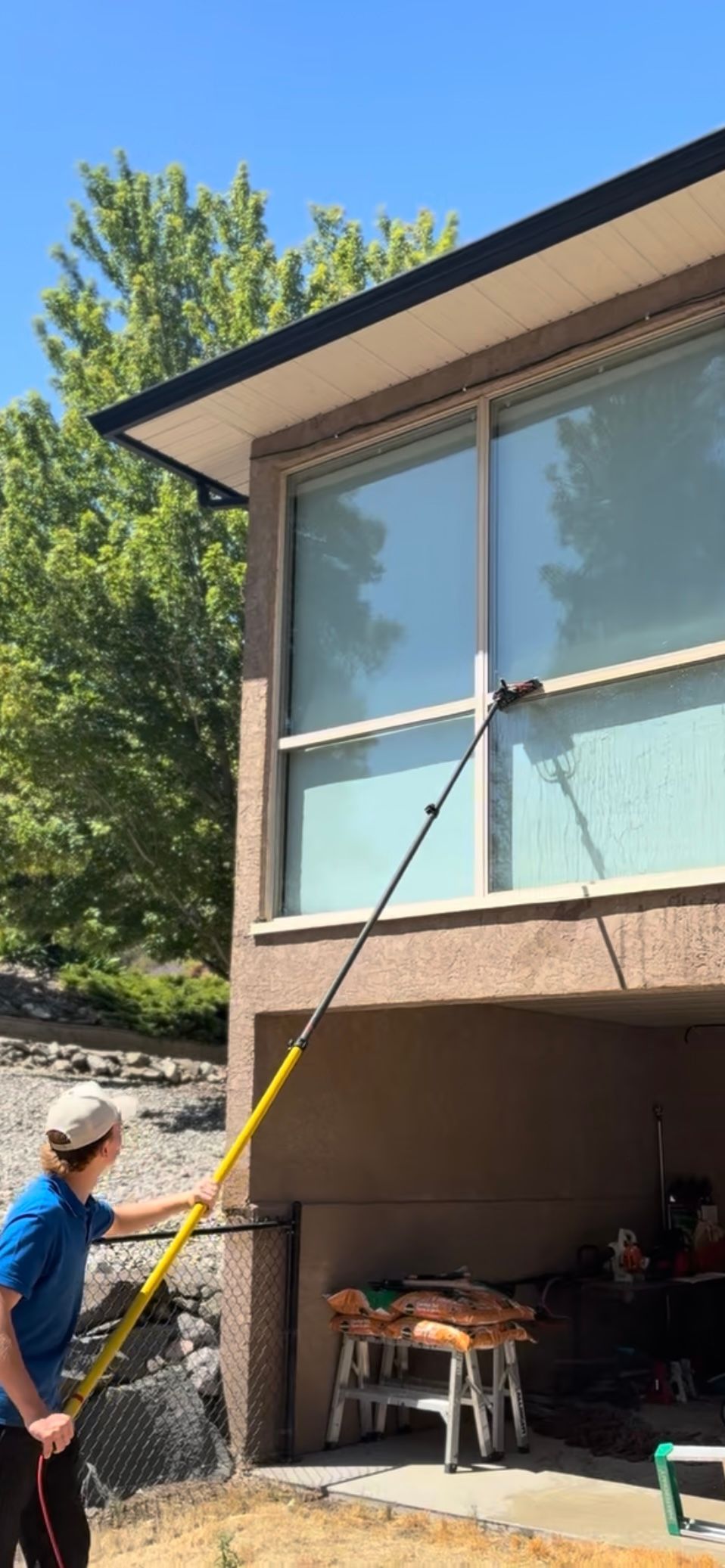 Person washes a window on a two-story building using a pole. The person is wearing a hat and blue shirt. Sunny day.