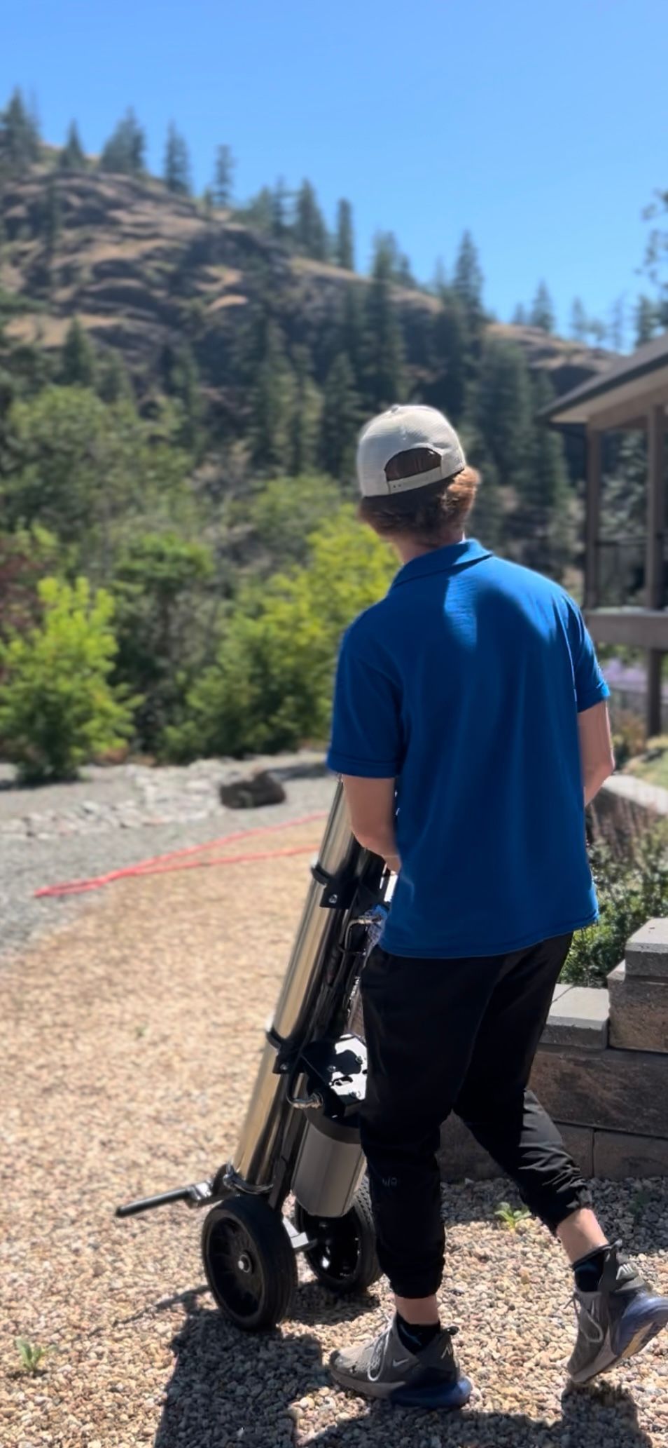 Person in blue shirt and hat walking with a golf push cart on a sunny day outdoors.