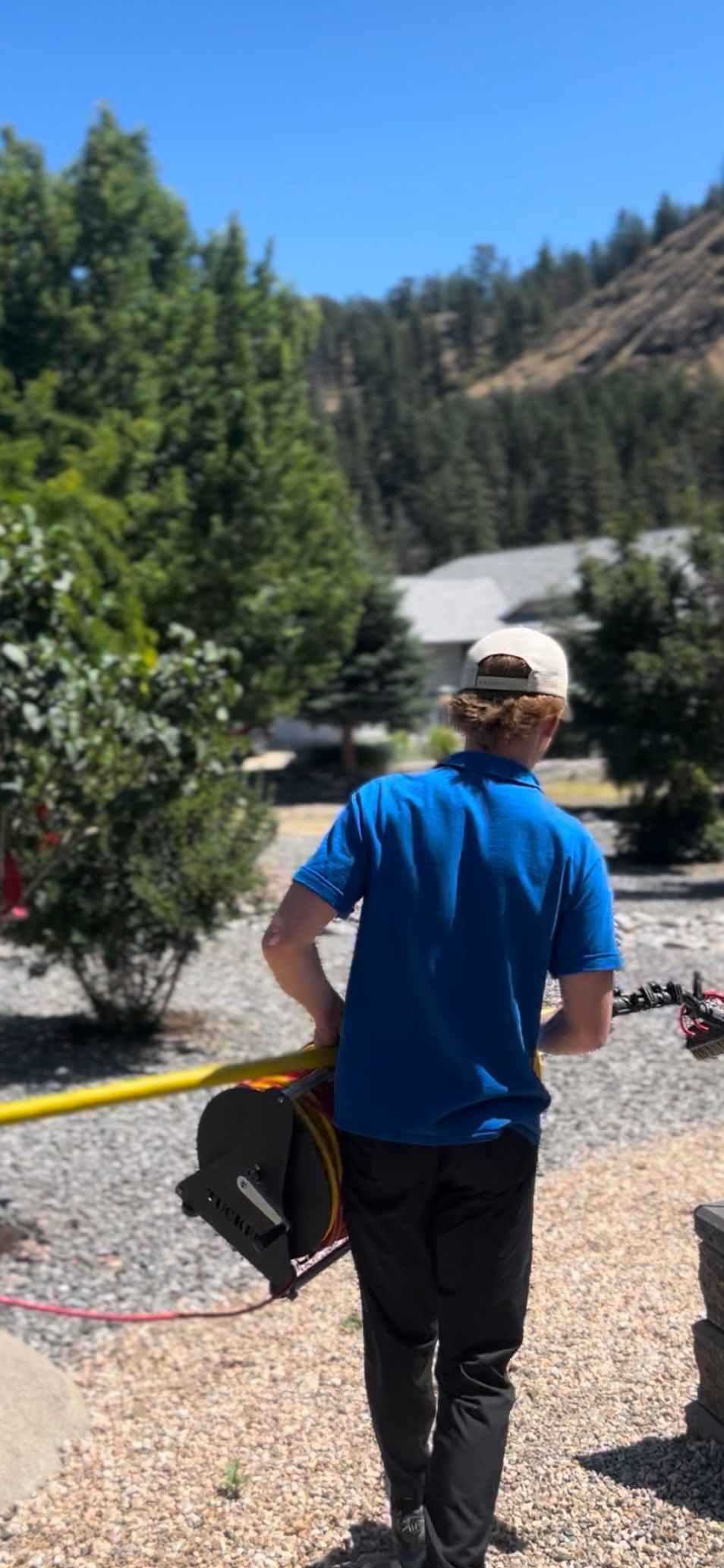 A person carrying equipment, walking towards the camera. Outdoors, bright blue sky and trees.