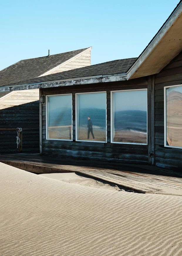 Weathered wooden building with large windows reflecting a person standing on a beach.