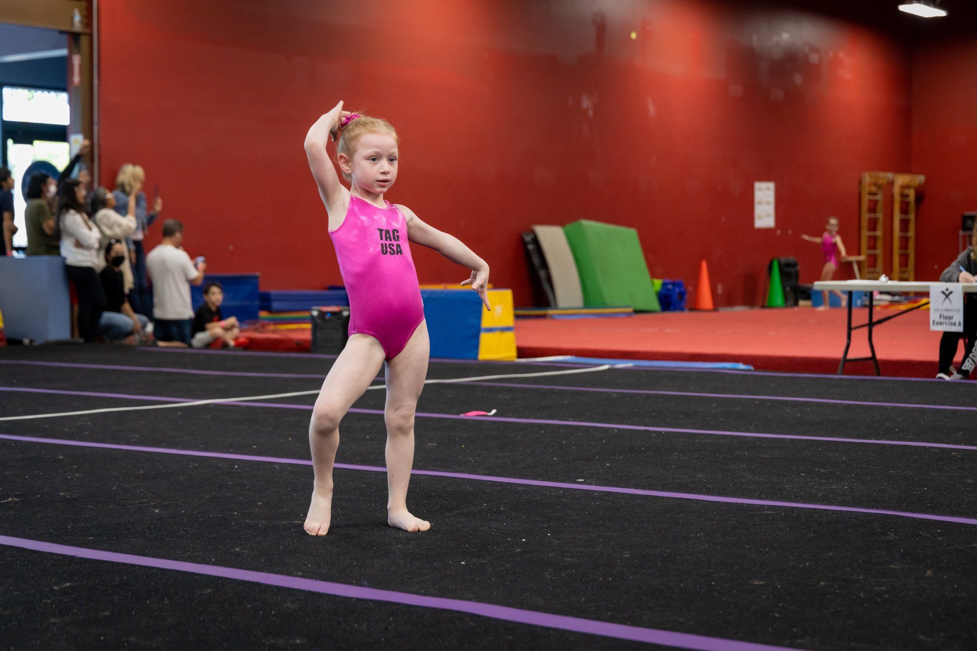 A little girl in a pink leotard is doing a balance beam on a gym floor.