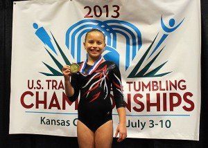 A young girl is holding a medal in front of a 2013 u.s. tumbling championships banner