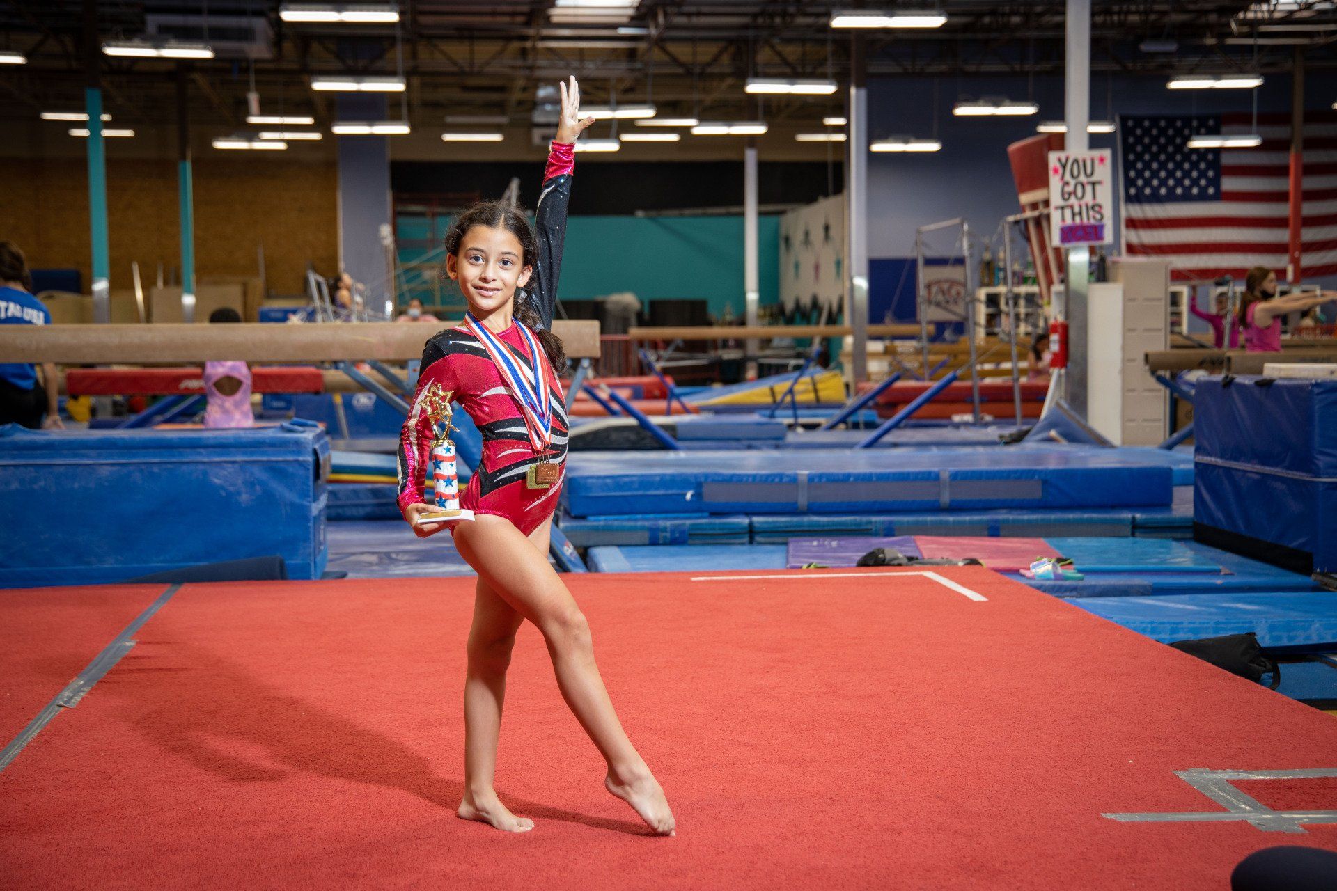 A young girl is standing on a balance beam in a gym holding a trophy.