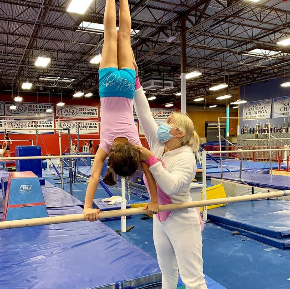 A woman is helping a young girl do a handstand on a bar in a gym.