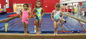 Three young girls are standing on a balance beam in a gym.