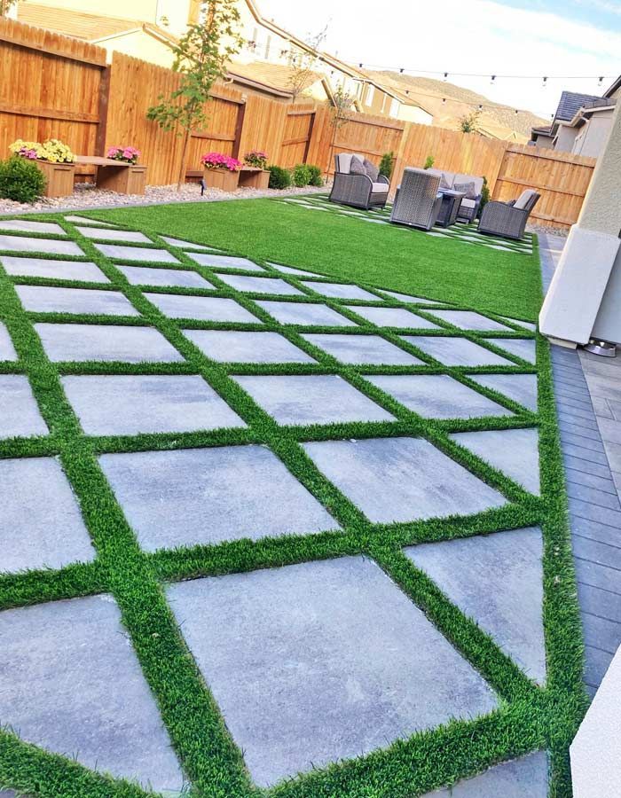 Concrete pavers with grass strips in a backyard. Wooden fence, seating area, and green lawn in the background.