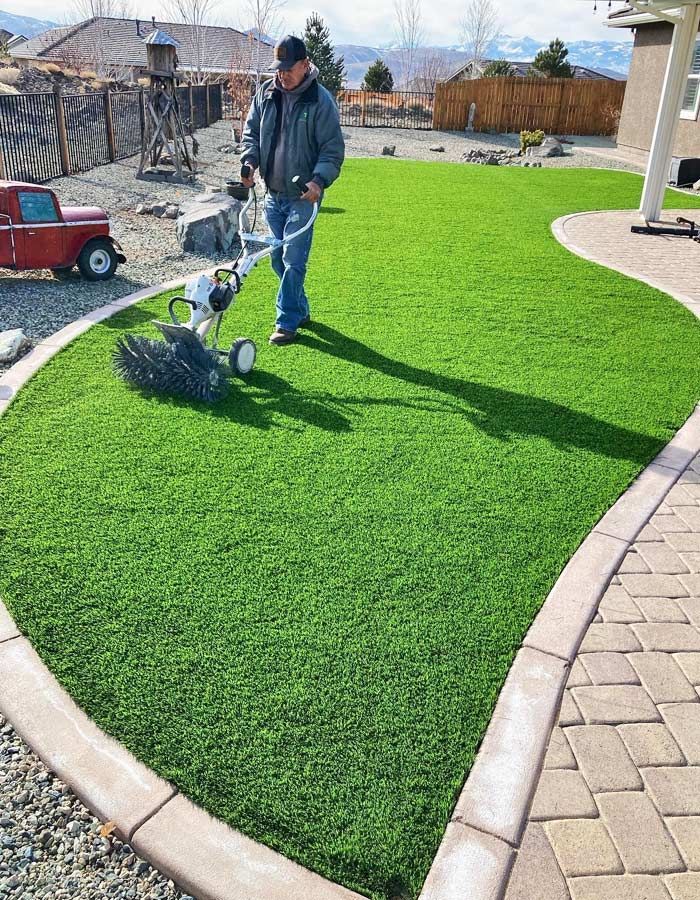 Man using a power brush on a bright green artificial lawn, bordered by a tan curb and patio.