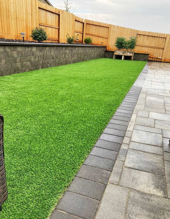 Lush green artificial lawn in a backyard, bordered by pavers and a retaining wall with a wooden fence.