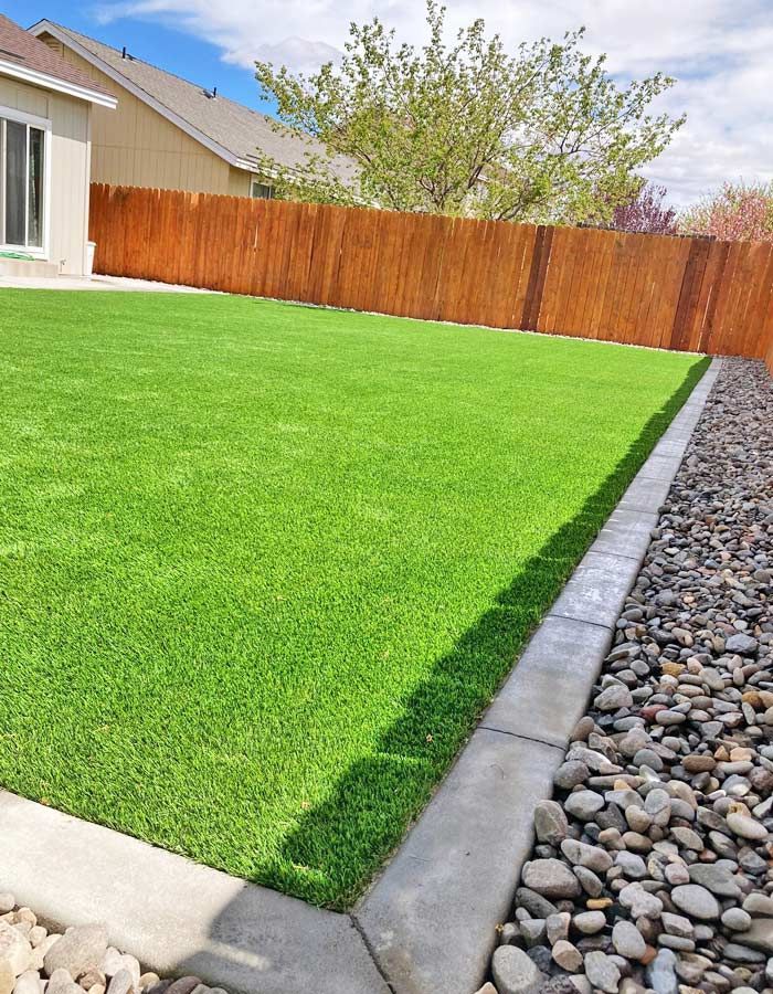 Green artificial turf lawn in a backyard, bordered by a concrete edge and rocks, with a wooden fence.