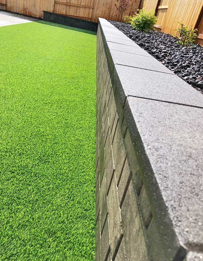 Green artificial turf lawn next to a grey brick retaining wall with black gravel above.