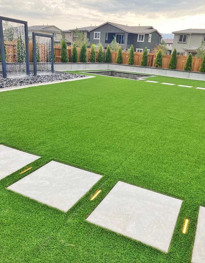 Backyard with artificial green turf, square stepping stones with lights, and a water feature.
