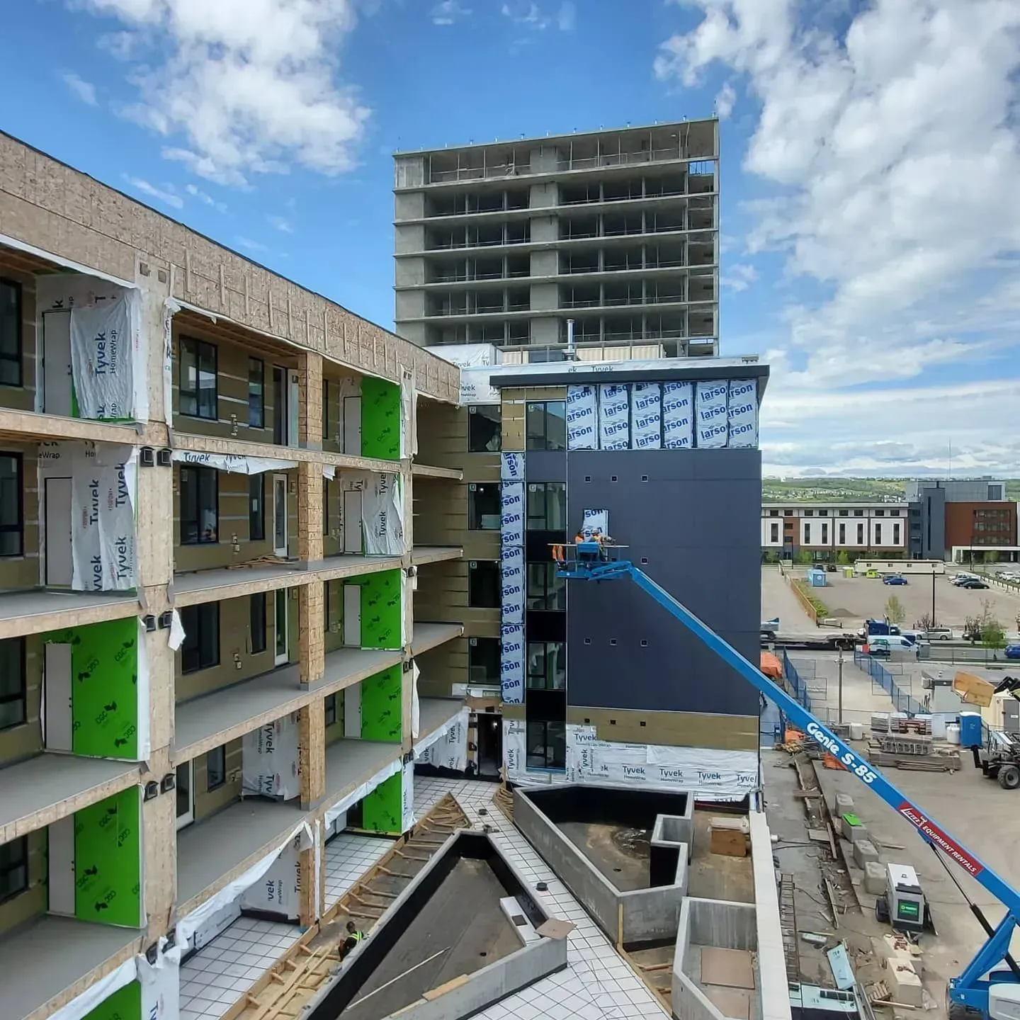 Construction site with unfinished multi-story buildings, blue crane boom, and green panel accents under a partly cloudy sky