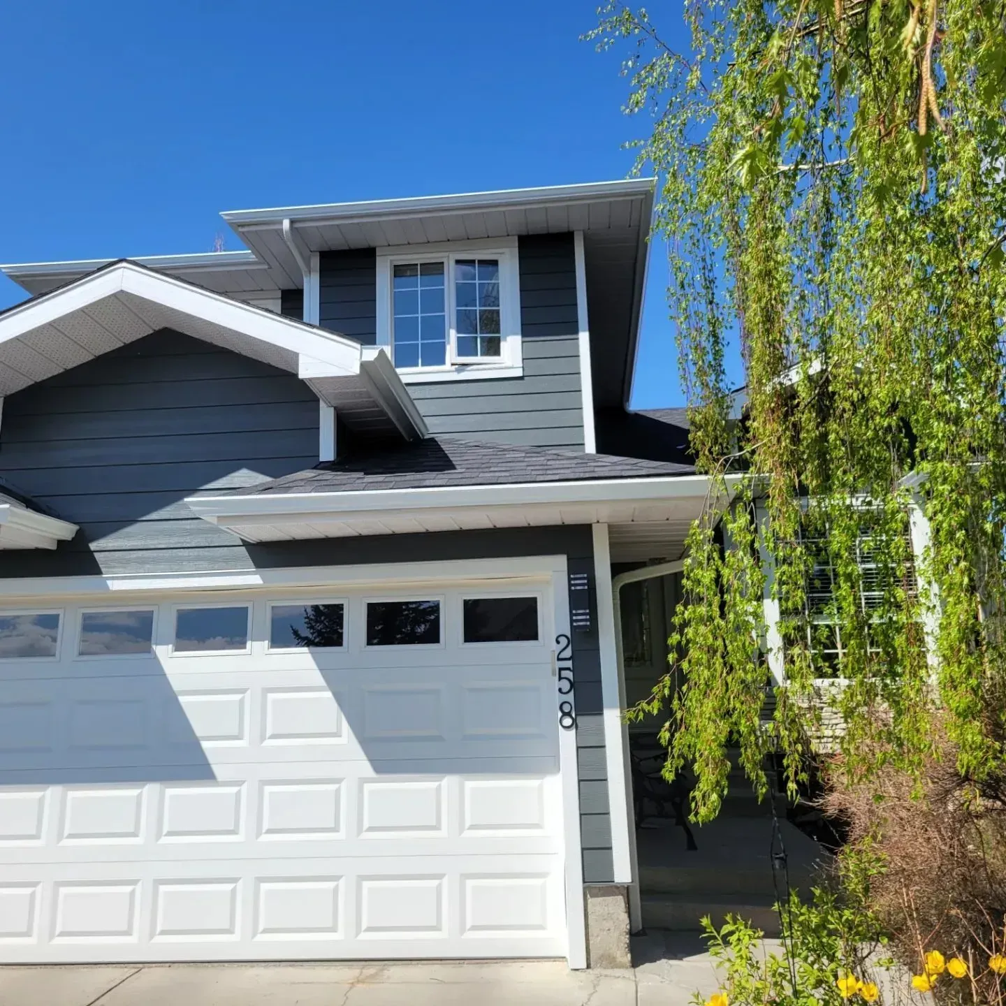 Blue-gray two-story house with white garage and willow tree in front yard