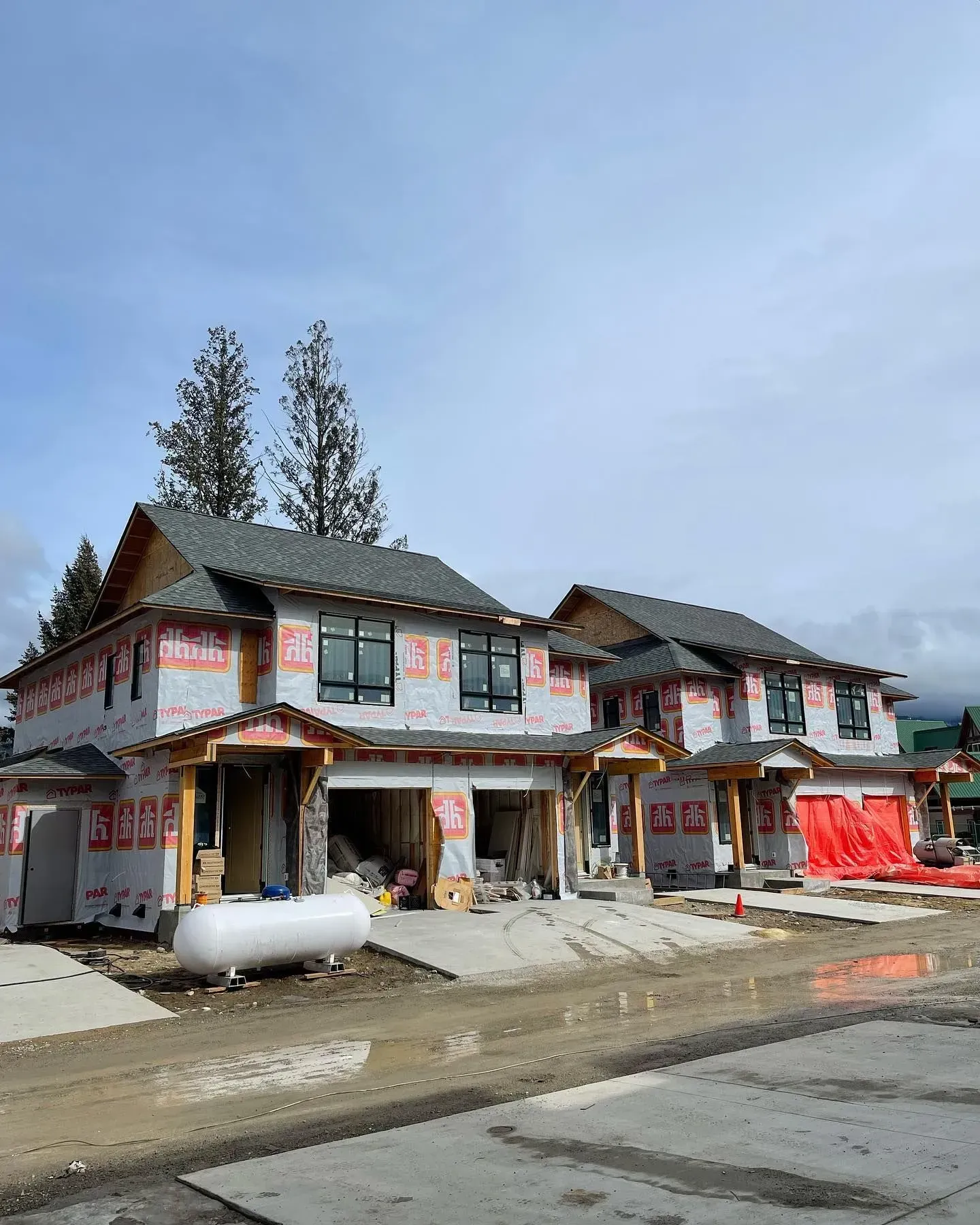 A row of unfinished buildings with white walls and red trim on a muddy roadside under a cloudy sky.