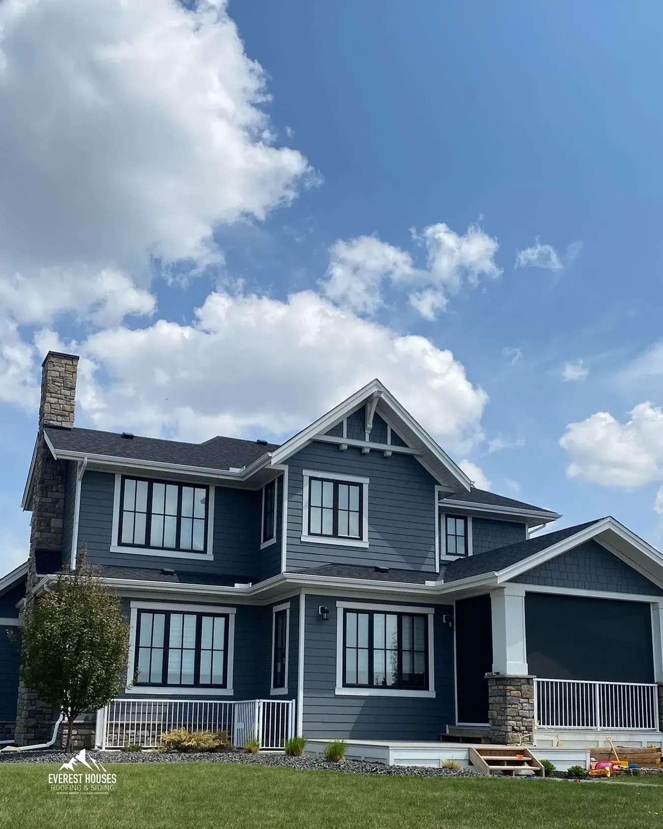 Blue-gray two-story house with white trim and dormers under a partly cloudy sky