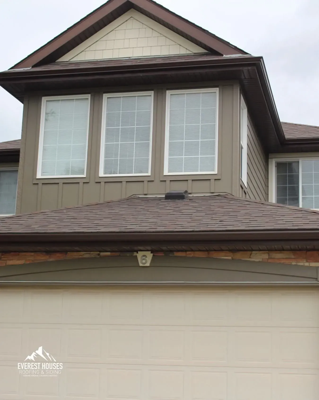Two-story beige house exterior with white windows and a brown shingled roof above a garage