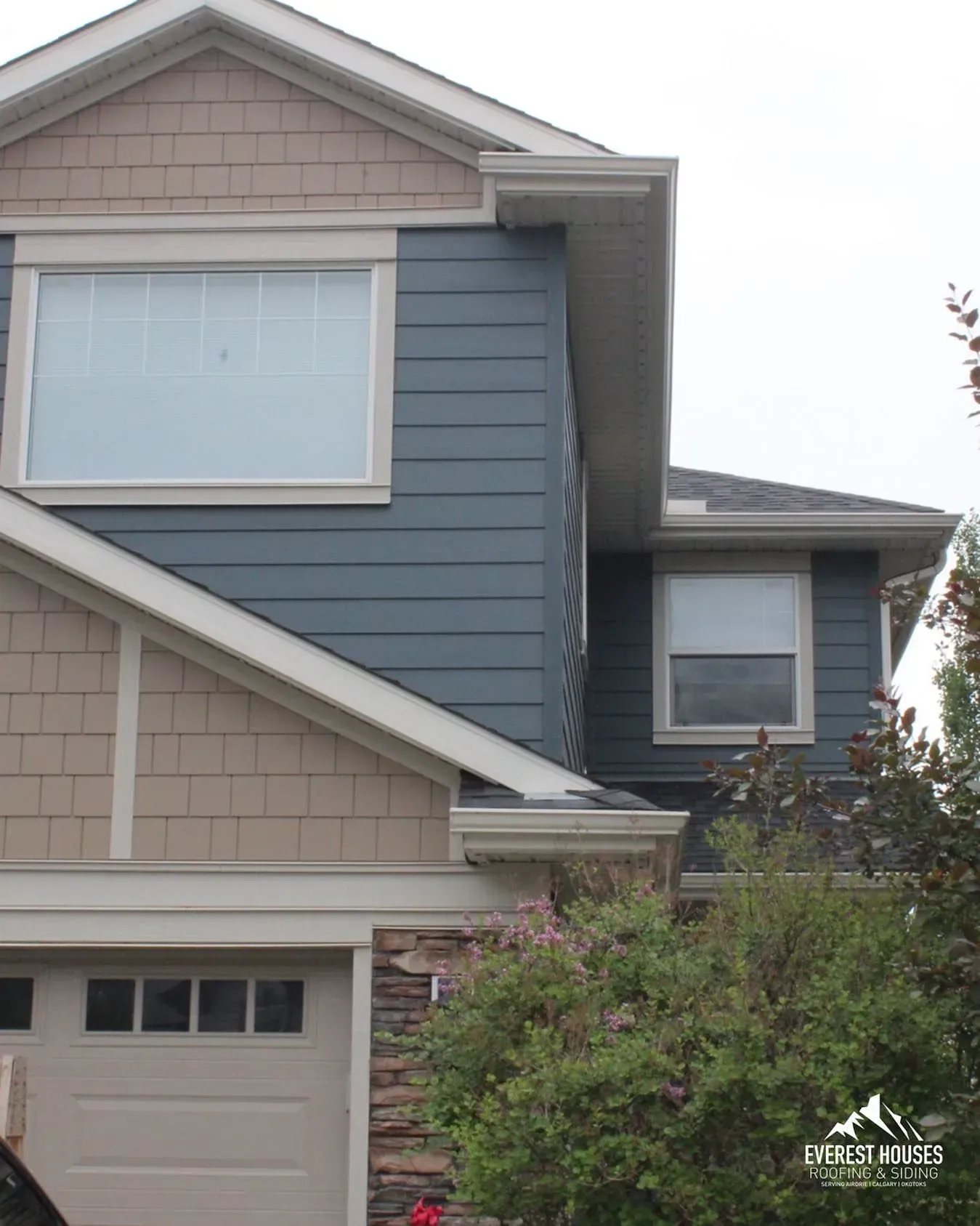 Two-story suburban house with blue-gray siding, tan shingles, and a garage, partly obscured by bushes.