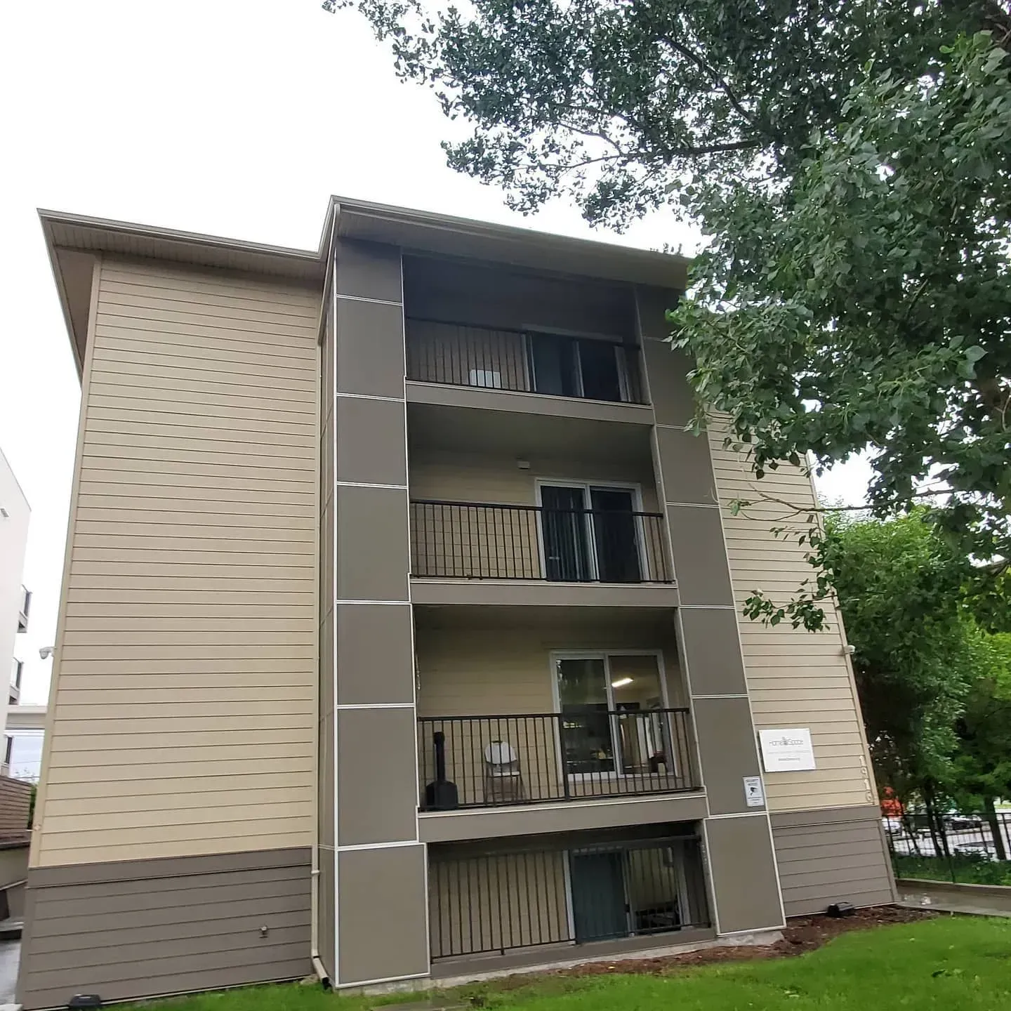Three-story beige apartment building with balconies, viewed from below beside trees
