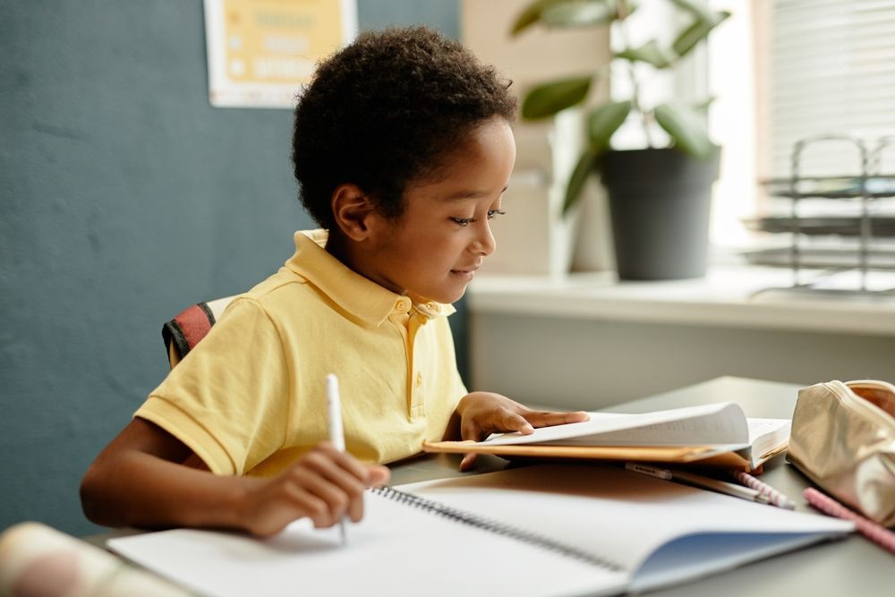 A child in a yellow shirt writing in a notebook at a desk.