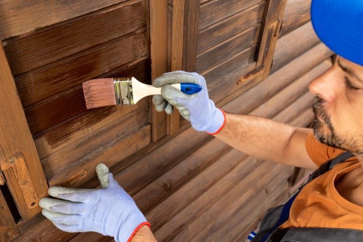 Person painting wooden shutters with a brush; wearing gloves, and a blue hard hat.