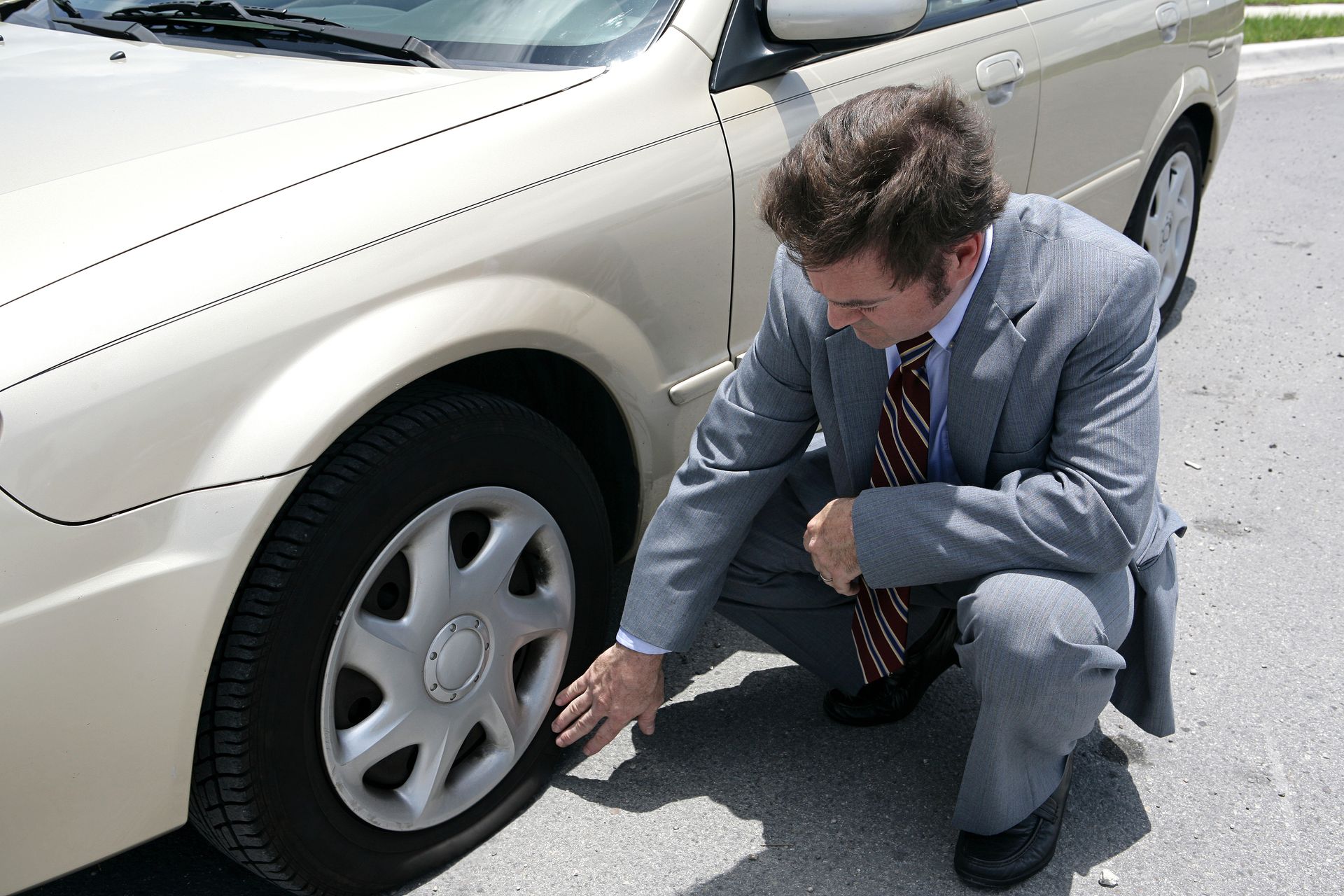 A man in a suit and tie is kneeling next to a car looking at the tire